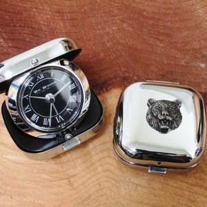 Könnte beinhalten: Eine silberne Taschenuhr mit schwarzem Zifferblatt und römischen Ziffern. Die Uhr ist geöffnet und das Zifferblatt ist sichtbar. Die Uhr befindet sich in einem silbernen Gehäuse mit einem gravierten Bären auf dem Deckel.