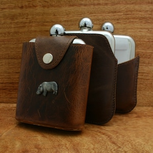 Puede incluir: Tres matraces de acero inoxidable con fundas de cuero marrón. El matraz de la izquierda tiene un pequeño emblema de rinoceronte de metal.