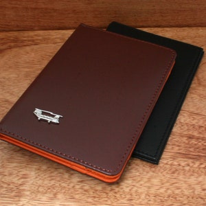 Puede incluir: Dos porta pasaportes de cuero, uno marrón con un borde naranja y un diseño de avión plateado, y otro negro.