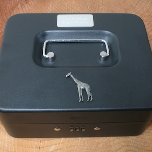 Puede incluir: Caja de metal negra con una cerradura de combinación y una figura de jirafa plateada en la tapa. La tapa tiene una manija plateada y una placa plateada que dice "Safari Collection".