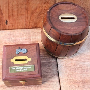 May include: Two wooden money boxes. One is a barrel shape with brass bands and a slot on the top. The other is a square box with a brass latch and a small tractor figurine on the top. The box has a brass plaque that says "Your Message Engraved Here For Free".