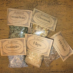 May include: Six small bags of dried herbs with brown paper labels. The labels have a decorative border and the text "The Herb Peddler" at the top. The herbs are labeled "Lemon Balm", "Chamomile", "Lavender", "Hops", "Holy Basil".