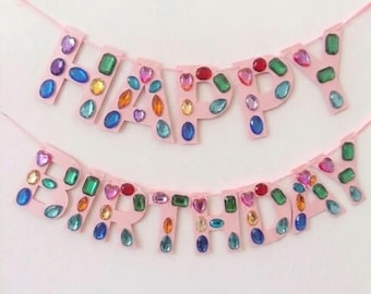 May include: A two-line banner spelling out HAPPY BIRTHDAY in light pink cardboard letters. Each letter is decorated with various colorful, faceted plastic gemstones in shapes including ovals, rectangles, and hearts in shades of blue, green, red, and orange.