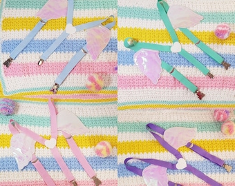 May include: Four pairs of colorful suspenders with iridescent angel wings. The suspenders are on a striped fabric background. The suspenders are in blue, green, pink, and purple.