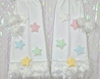 May include: Two white decorative items, each with pastel-coloured star embellishments and a fluffy white trim. They are tied at the top with white ribbon and have fluffy white accents. The background is a shimmering, iridescent material.