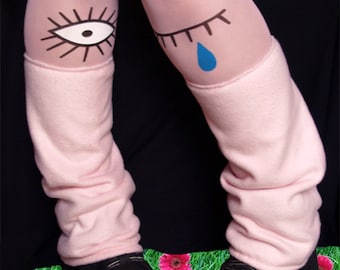 Può includere: Calze rosa con un occhio nero e un occhio nero con una lacrima blu disegnata su di esse. Le calze sono indossate con scarpe da piattaforma nere e la persona è in piedi su un letto di erba verde con fiori rosa.