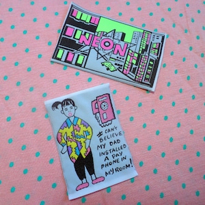 May include: Two iron-on patches. The first patch is a white background with a neon green and black cityscape with the word "NEON" in pink. The second patch is a white background with a cartoon illustration of a person with a pink phone and the text "I CAN'T BELIEVE MY DAD INSTALLED A PAY PHONE IN MY ROOM!"