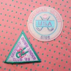 May include: Two embroidered patches. One is a white circle with pink and blue embroidery that says "Professional Golfers USA Side". The other is a green triangle with pink and white embroidery that says "Country Club Down Under '88".