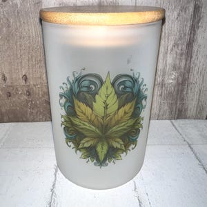 May include: Frosted glass candle with a bamboo lid. The candle features a green and yellow cannabis leaf design with blue and green swirls. The candle is cylindrical and sits on a white brick surface.