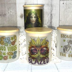 May include: Four frosted glass jars with wooden lids. The jars feature various designs, including a woman's portrait, the words "Good Vibes", a cannabis leaf with sunglasses and the word "Blazed", and a skeleton with the words "Stoned to the Bone".