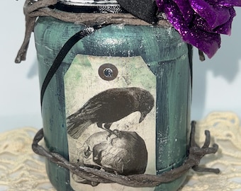 Può includere: Un barattolo dipinto di verde acqua con l'immagine di un corvo nero e un teschio su un'etichetta. Il barattolo è decorato con una rosa glitterata viola, un nastro nero e abbellimenti simili a ramoscelli. Il barattolo è su una superficie crema a trama aperta.