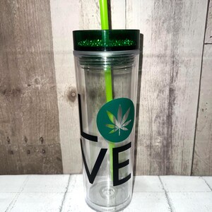 May include: Clear plastic tumbler with a green lid and straw. The word "LOVE" is printed vertically in black, with a green circle and cannabis leaf design. The lid has green glitter.