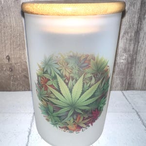 May include: A frosted glass candle holder with a wooden lid. The candle holder features a colorful cannabis leaf design. The design is a mix of green, red, and yellow leaves. The candle holder is on a white surface.