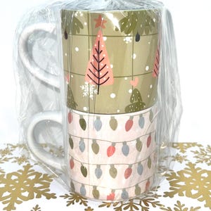 May include: Two white-handled ceramic mugs stacked and wrapped in clear cellophane. The top mug features a pink Christmas tree design on a tan background. The bottom mug has a string of Christmas lights design on a pink background.