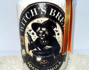 Può includere: Un barattolo bianco con finitura anticata, con un'etichetta in bianco e nero che recita "Witch's Brew Toxic Formula". L'etichetta raffigura una strega che mescola un calderone. Il barattolo è adornato con nastro arancione e nero e un coperchio decorativo.