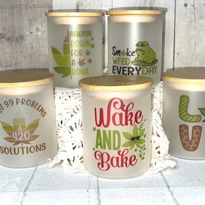 Puede incluir: Cinco frascos de vidrio esmerilado con tapas de madera. Cada frasco tiene un diseño diferente con texto y gráficos de hojas de cannabis. El texto incluye "I got 99 problems & 420 solutions", "Always down for a bowl", "Smoke weed everyday", "Wake and Bake" y "Love".
