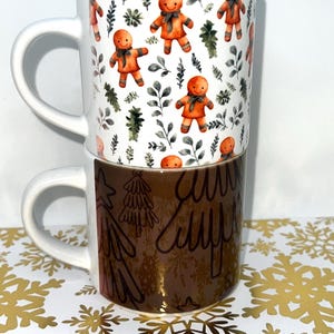 May include: Two stacked ceramic mugs. The top mug features a white background with gingerbread men and green foliage. The bottom mug has a brown metallic finish with gold tree designs. Both mugs have white handles.