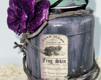 Può includere: Un barattolo decorativo con un'etichetta che recita "Frog Skin". Il barattolo è dipinto di grigio e viola, adornato con un fiore viola glitterato e avvolto con finte viti. Il coperchio è dipinto con accenti verdi e neri.