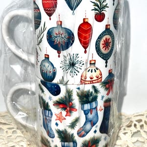 May include: Two white ceramic mugs stacked together, wrapped in clear cellophane. The mugs are decorated with Christmas ornaments, stockings, and greenery in red, blue, and green. Each mug has a white handle.