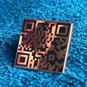 Puede incluir: Un pin de metal plateado con un diseño de código QR en blanco y negro.