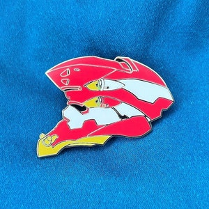 May include: Red, white, and yellow enamel pin of a robot head. The robot head is in a 3D style and has a detailed design.