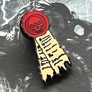 Może przedstawiać: Szpilka emaliowana w kolorze czerwonym i czarnym z czaszką i skrzyżowanymi kośćmi w projekcie pieczęci woskowej. Pieczęć jest przymocowana do żółtej wstążki z czerwonym napisem.