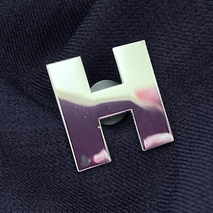 May include: A polished silver-tone metal lapel pin shaped like the capital letter H. The surface of the letter is highly reflective, showing distorted light reflections. The pin is attached to a dark navy blue fabric with a fine knit texture.