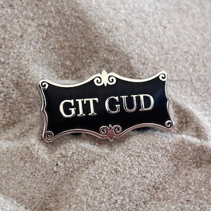Op de afbeelding: Een zwarte en zilveren emaille pin met de tekst "GIT GUD" op een witte zand achtergrond.