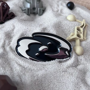 May include: A silver and black enamel pin with a swirl design. The pin is surrounded by small toy building blocks on a sandy surface.