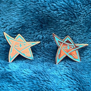 May include: Two gold-coloured enamel pins with a five-pointed star design. The stars are outlined in turquoise blue and have a red interior.