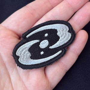 May include: Black embroidered patch with a stylized design featuring a central circle and two curved, wing-like shapes in light gray. The patch is oval-shaped and has a black border.