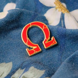 Könnte beinhalten: Eine Anstecknadel aus goldfarbenem Metall in Form des griechischen Buchstabens Omega. Die Nadel ist mit leuchtend rotem Emaille gefüllt, das eine dezente, marmorierte Textur aufweist und einen Kontrast zu den goldfarbenen Metallrändern bildet.