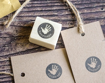 May include: A set of two brown paper gift tags with a black handprint and the word "handmade" printed on them. The tags are tied with twine and are next to a wooden block stamp with the same design.