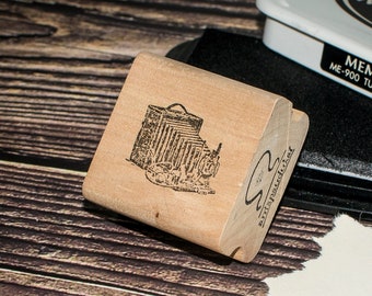 May include: A wooden stamp featuring a black and white image of a vintage camera. The stamp is on a wooden surface. The text on the stamp reads "Memento Tuxedo Black".