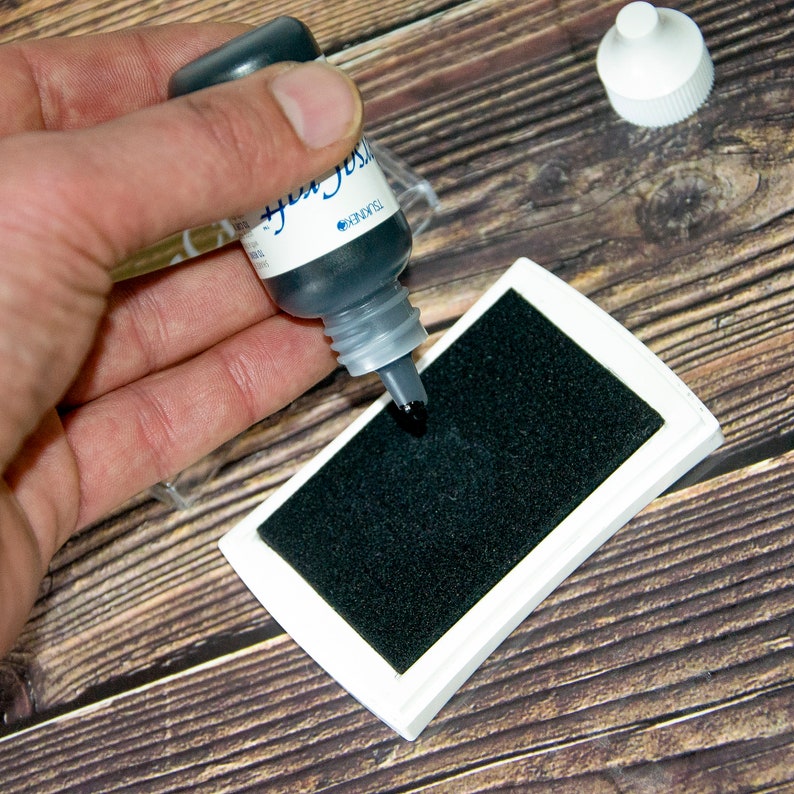 K&ouml;nnte beinhalten: Eine Hand h&auml;lt eine kleine Flasche mit schwarzer Tinte mit der Aufschrift "Black Soot" &uuml;ber einem wei&szlig;en Stempelkissen mit einem schwarzen Stempelkissen.