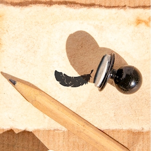 Pode incluir: Um carimbo de borracha preto com um desenho de pena sobre um fundo branco. O carimbo está sobre um pedaço de papel castanho com um lápis ao lado.
