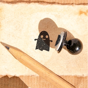 Op de afbeelding: Een zwarte rubberen stempel met een spookontwerp. De stempel ligt op een stuk bruin papier met een potlood ernaast.