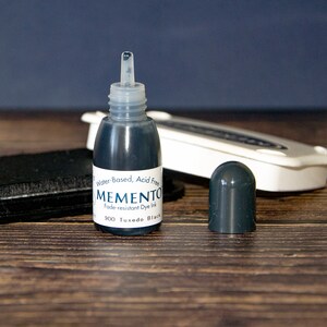 May include: A small bottle of black ink labeled "MEMENTO Fade-resistant Dye Ink 900 Tuxedo Black" with a clear dropper lid. The bottle is labeled "Water-Based, Acid Free".