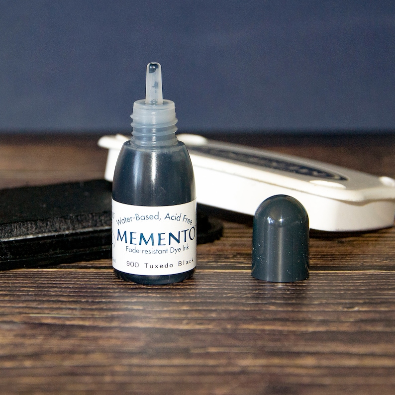 May include: A small bottle of black ink labeled "MEMENTO Fade-resistant Dye Ink 900 Tuxedo Black" with a clear dropper lid. The bottle is labeled "Water-Based, Acid Free".