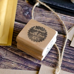 May include: A wooden stamp with the words "handmade with love 100%" in a circular design. The stamp is light brown and sits on a wooden surface with twine and craft paper.