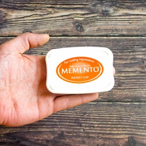 May include: A white ink pad with the text "For Lasting Impressions Fade-resistant Dye Ink MEMENTO POTTER'S CLAY" in orange.