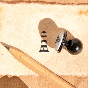 May include: A black and white rubber stamp with a lighthouse design. The stamp is on a piece of brown paper with a pencil lying on top of it.