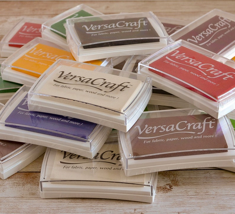 May include: A stack of ink pads in various colors, each with a clear plastic lid and the text "VersaCraft" printed on the top. The ink pads are labeled "For fabric, paper, wood and more!"