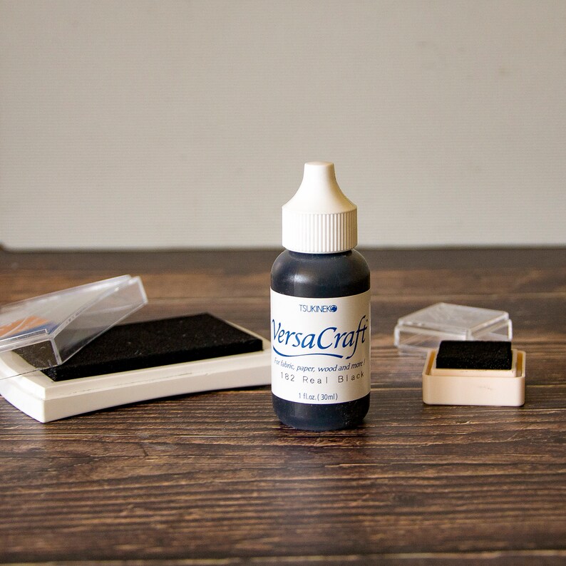 K&ouml;nnte beinhalten: Eine Flasche schwarzer VersaCraft-Tinte f&uuml;r Stoff, Papier, Holz und mehr. Die Flasche ist mit "182 Real Black" und "1 fl oz (30ml)" beschriftet.