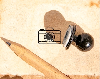 May include: A black rubber stamp with a camera icon on a brown paper background. A wooden pencil is lying on the paper.