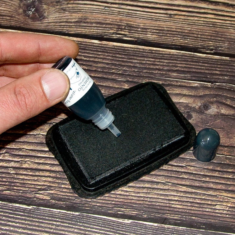 May include: A black ink pad with a clear bottle of black ink being squeezed onto the pad. The bottle has the text "Dye-based Ink" and "Memento" on it.
