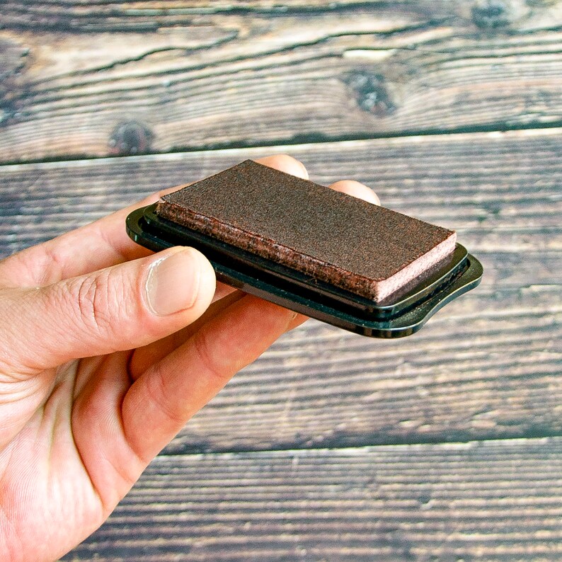 May include: A brown ink pad in a black plastic case. The ink pad is used for stamping.
