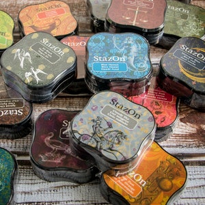 May include: A collection of StazOn brand ink pads in various colors and designs. The ink pads are square shaped with rounded corners and feature different patterns and illustrations. The ink pads are arranged in a pile on a wooden surface.
