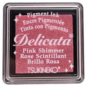 May include: A pink ink pad with the text "Delicata" in a cursive font. The ink pad is labeled "Pink Shimmer, Rose Scintillant, Brillo Rosa" and "Pigment Ink, Encre Pigment&eacute;e, Tinta con Pigmento". The ink pad is made by TSUKINEKO.
