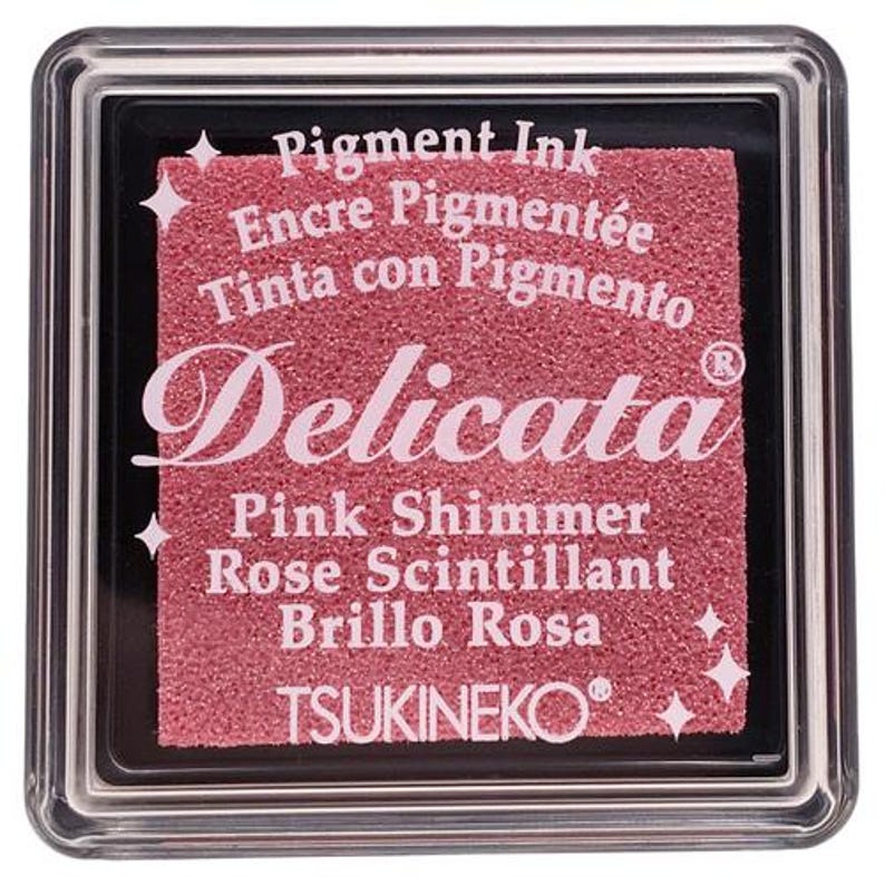 May include: A pink ink pad with the text "Delicata" in a cursive font. The ink pad is labeled "Pink Shimmer, Rose Scintillant, Brillo Rosa" and "Pigment Ink, Encre Pigment&eacute;e, Tinta con Pigmento". The ink pad is made by TSUKINEKO.
